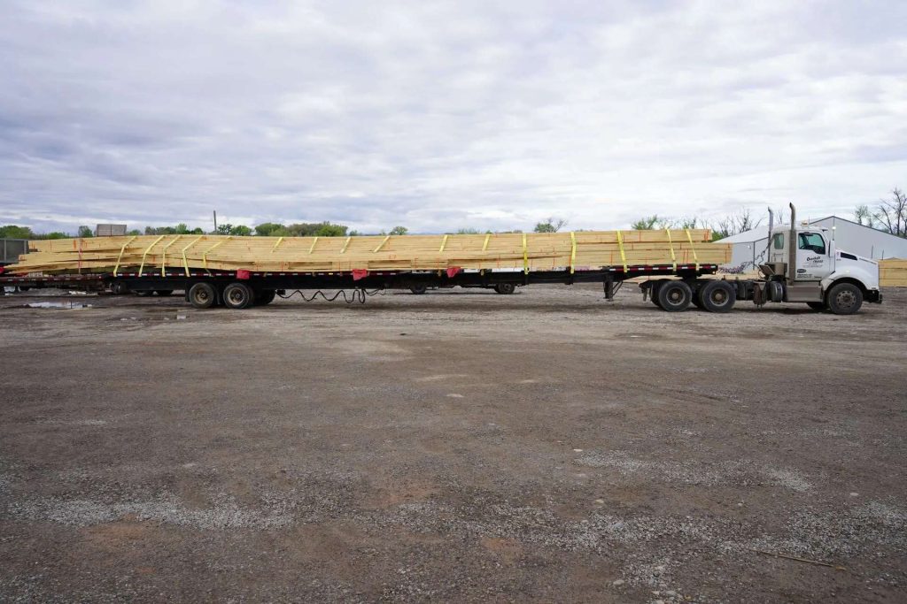 A flatbed semi-truck is parked on a gravel lot, carrying a long load of wooden boards secured with yellow straps. The sky is overcast, and trees and buildings are visible in the background.