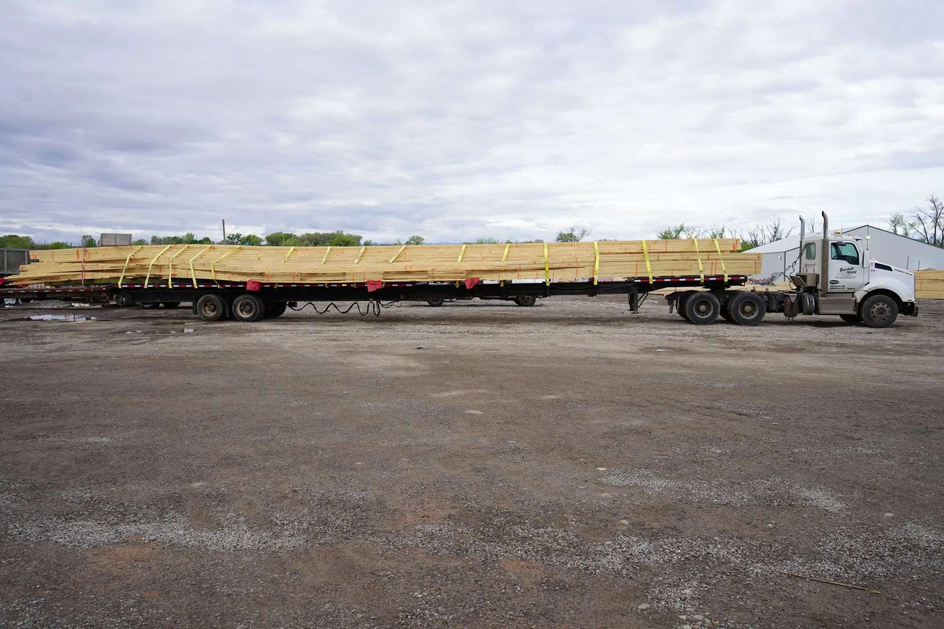 A flatbed semi-truck is parked on a gravel lot, carrying a long load of wooden boards secured with yellow straps. The sky is overcast, and trees and buildings are visible in the background.