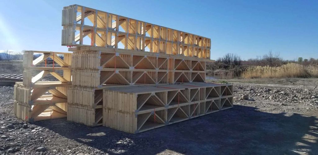 Stacks of large wooden trusses are arranged on bare ground at a construction site, with dry grass, shrubs, and a clear blue sky in the background.