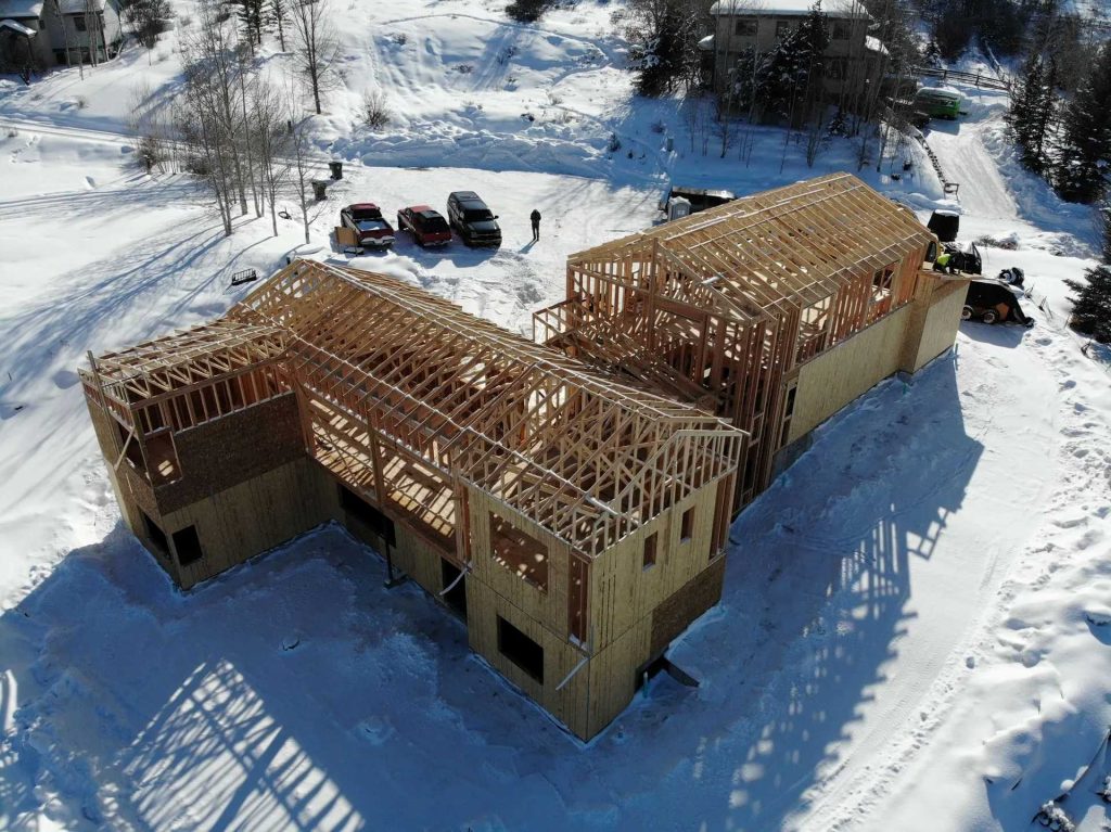 Aerial view of a wooden house under construction in a snowy landscape, with vehicles and people nearby. The frame and roof trusses are visible, and snow covers the surrounding ground.